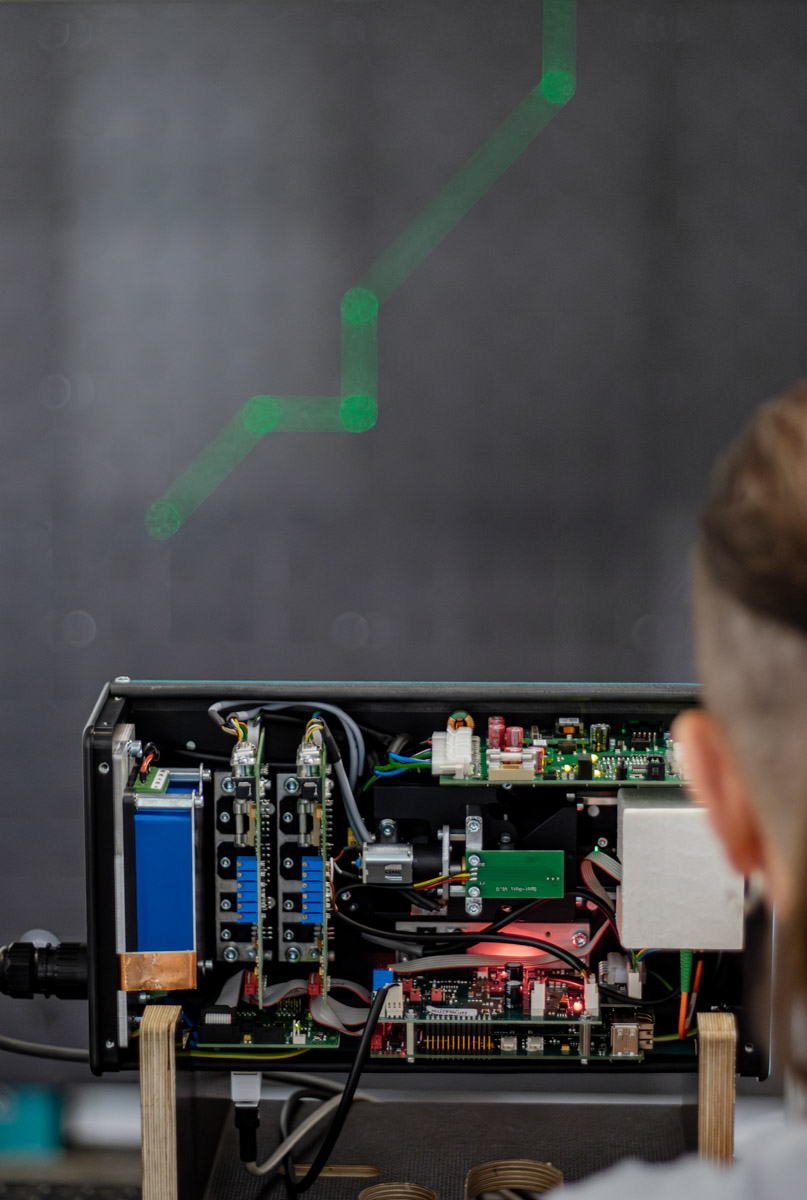 Geöffneter SL-Laser Projektor mit sichtbaren elektronischen Komponenten – Techniker überprüft die Bauteile, während grüne Laserlinien projiziert werden.