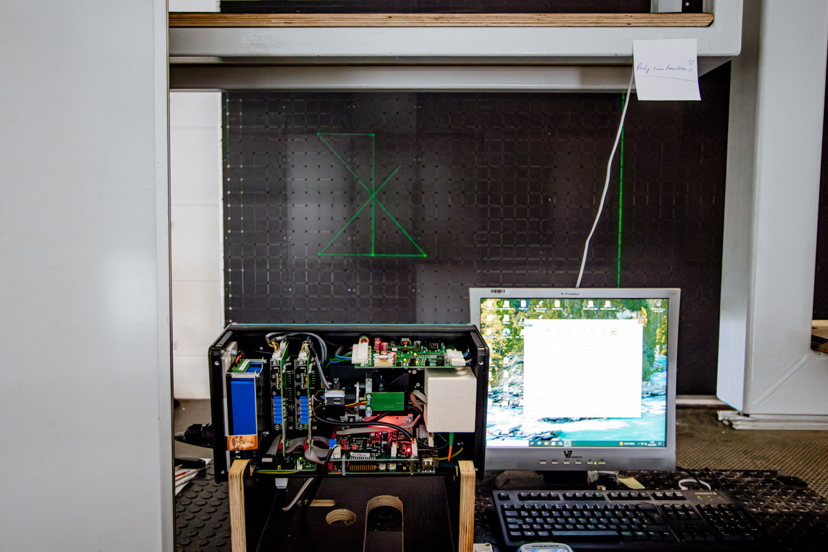 Geöffnetes SL-Lasergerät mit angeschlossenem Computer, das für Tests und Kalibrierung auf einer schwarzen Kalibrierungsfläche grüne Laserlinien projiziert.