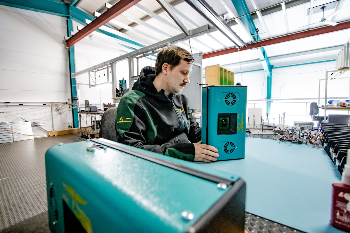 Mitarbeiter in SL-Laser-Werkstatt prüft ein grünes SL-Lasergerät auf einem Arbeitstisch in moderner Produktionsumgebung.