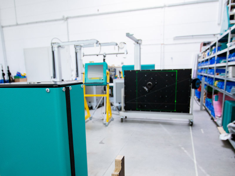SL-Lasergerät in einer Fertigungshalle, ausgerichtet auf eine schwarze Kalibrier- und Projektionsfläche mit grünem Laserbild, im Hintergrund weitere Prüf- und Teststationen.