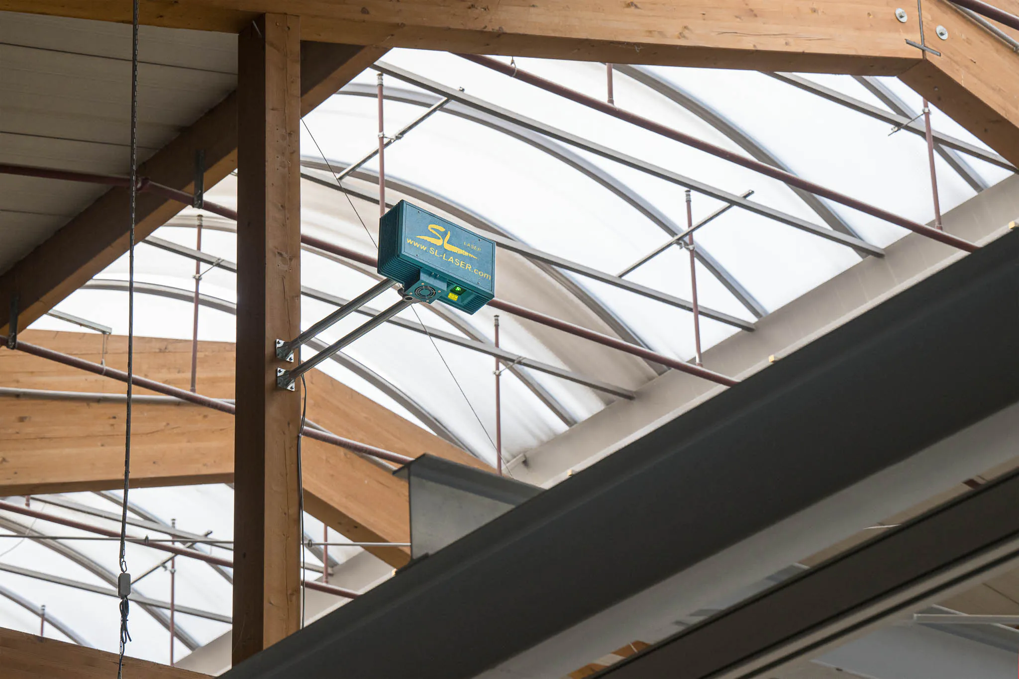 SL-Lasergerät ist an einer Holzkonstruktion unter dem Hallendach montiert und für präzise Laserprojektionen ausgerichtet.